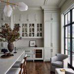 Kitchen with marble island, light gray cabinetry, wine fridge, and tall black-framed windows.