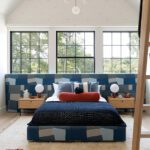 Modern boy’s bedroom with blue upholstered headboard, red accent chandelier, and large black-framed windows bringing in natural light.