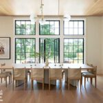 Expansive dining area bathed in natural light with pale wood flooring, a long minimalist table, modern brass lighting, and wall-to-wall windows.