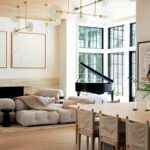The airy great room combines a sculptural dining table and plush seating with modern brass lighting and expansive windows framing garden views.