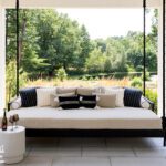 A serene covered patio features a cushioned swing daybed surrounded by lush greenery and views of a pond beyond.