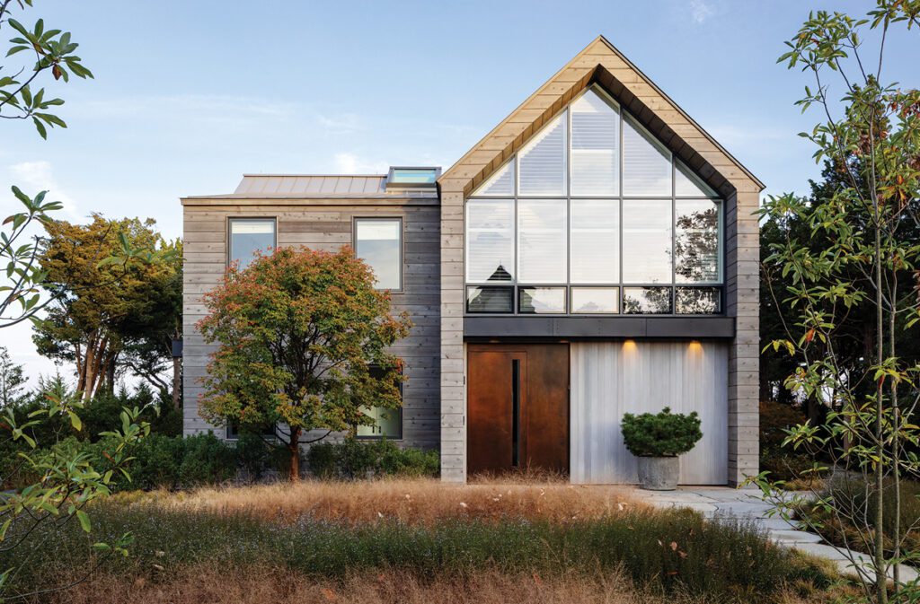 A modern exterior balances natural textures—stone, wood, and metal—with a dramatic glass gable that invites light into the home’s interior. A mature landscape of wild grasses softens the clean architectural lines.