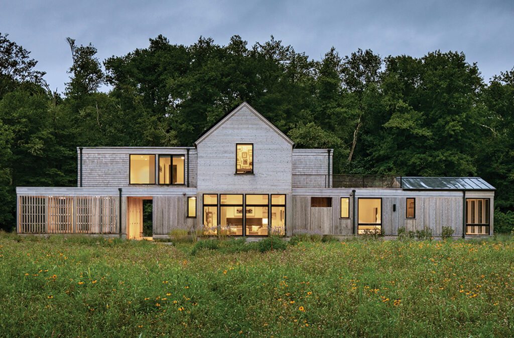 Modern farmhouse-style home with wood siding at twilight, set in a meadow with forest backdrop.
