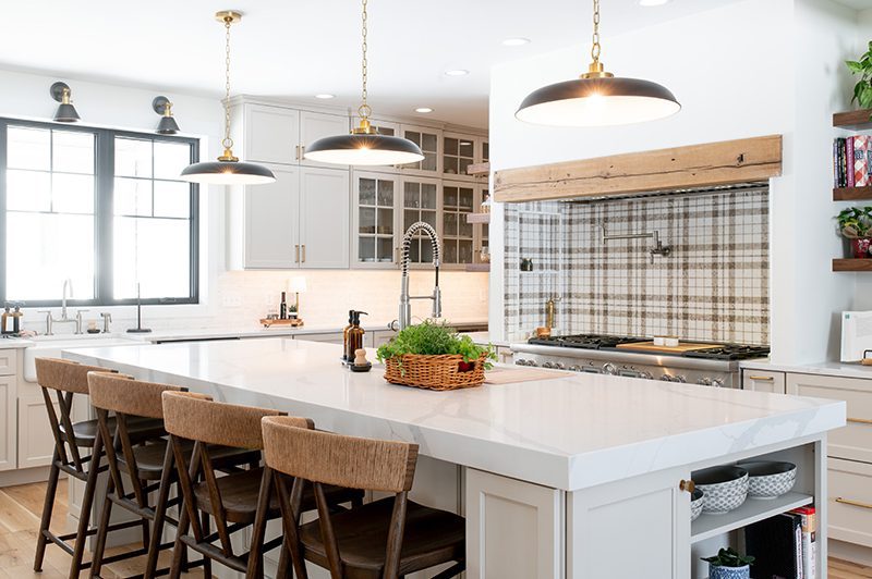 White kitchen with oversized island, plaid backsplash, brass accents, and pendant lighting.