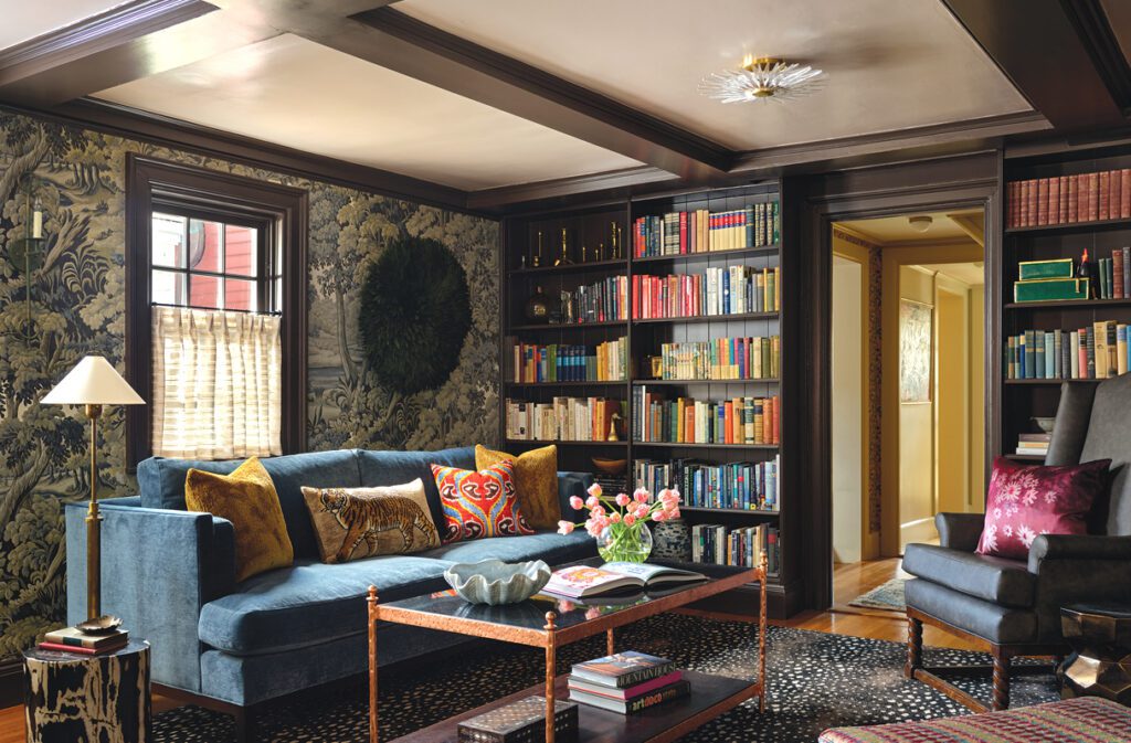 Moody home library with scenic wallpaper, built-in bookshelves, blue sofa, patterned rug, and dark wood trim.