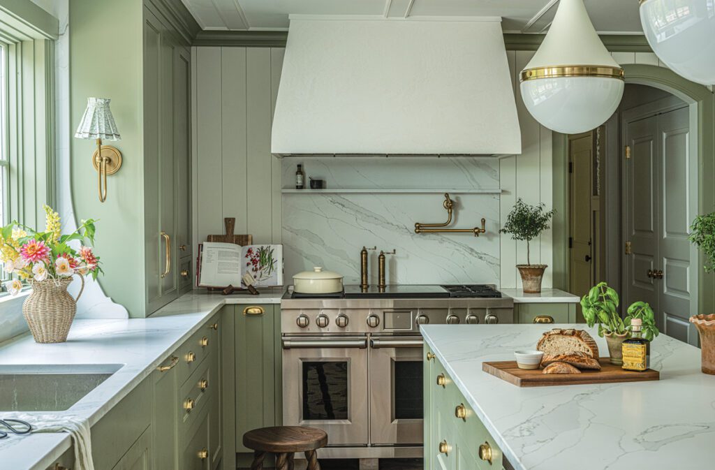 Bright kitchen breakfast nook with round wooden table, wicker chairs, sage green cabinetry, and pleated pendant light.